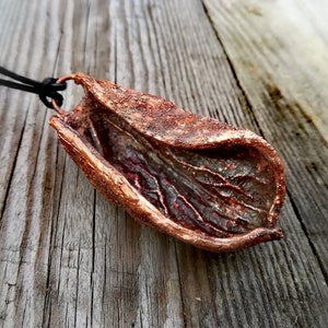 Puede incluir: Un collar con un colgante de hoja de color cobre y un cordón negro. El colgante está hecho de metal y tiene una superficie texturizada detallada.