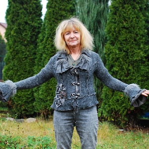 Puede incluir: Una mujer con cabello rubio lleva una chaqueta de punto gris con cuello de volantes y adornos decorativos. Está de pie frente a una fila de árboles de hoja perenne.