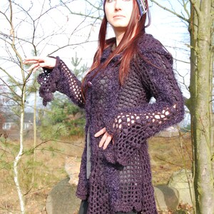 Può includere: Un cardigan di uncinetto viola con un design lungo e fluido. Il cardigan ha un motivo testurizzato, a trama aperta e un colletto largo e fluido. La modella indossa una fascia per capelli a motivi.