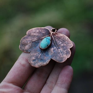 Puede incluir: Un colgante de hoja de cobre con una piedra turquesa engastada en el centro. La hoja está detallada con venas y tiene un aspecto rústico y envejecido.