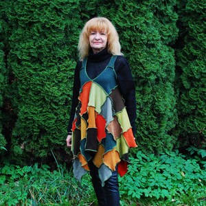 May include: A woman wearing a colourful patchwork dress with a black turtleneck sweater. The dress has a variety of colours and textures, including green, orange, brown, and blue. The woman is standing in front of a green hedge.