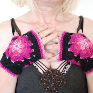 Puede incluir: Un par de guantes sin dedos de ganchillo negro y rosa con un diseño floral. Los guantes son usados por una persona con una blusa negra y encaje marrón.