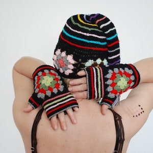 Puede incluir: Un gorro negro de ganchillo con rayas de colores y un diseño de flores. El gorro lo lleva una persona con los brazos en alto, llevando guantes sin dedos de ganchillo negros y de colores.