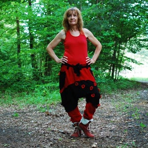 May include: A woman wearing a red tank top and black trousers with red accents stands in a forest. The trousers have a unique design with red circles and a red waistband.