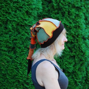 May include: A patchwork hat in shades of green, yellow, and blue, with a black brim and trim. The hat has a long, pointed tail in orange, brown, and black. The hat is worn by a person. The hat's design is reminiscent of a jester's cap.