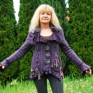 Può includere: Una donna indossa un cardigan di maglia viola con decorazioni e pantaloni neri, in piedi in un campo di erba verde con alberi sullo sfondo.