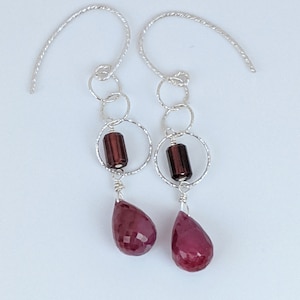 May include: A pair of silver dangle earrings with a teardrop-shaped red gemstone, a small brown cylinder bead, and multiple silver hoops.