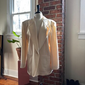 May include: A cream-colored blazer displayed on a mannequin. The blazer has a notched lapel, two front pockets, and a single-button closure. The garment is made of a soft, textured fabric. The mannequin is on a black stand, set against a brick wall.