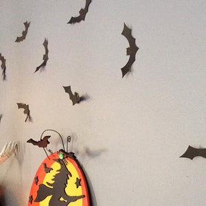 Op de afbeelding: Halloween-wanddecoratie met talrijke bruine vleermuisuitsparingen op een grijze muur. Een ketel met een skelet hand en een bord met een heksenthema zijn ook zichtbaar, wat een spookachtige sfeer cre&euml;ert.