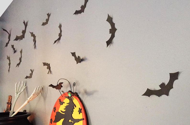 Op de afbeelding: Halloween-wanddecoratie met talrijke bruine vleermuisuitsparingen op een grijze muur. Een ketel met een skelet hand en een bord met een heksenthema zijn ook zichtbaar, wat een spookachtige sfeer cre&euml;ert.