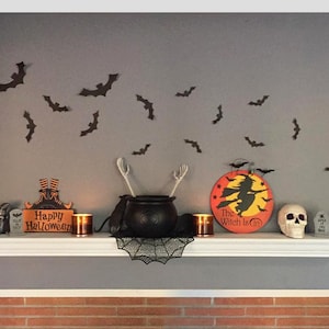 Op de afbeelding: Halloween-thema plankdecoratie met een ketel, schedel en vleermuizen. Inclusief een bord met "Happy Halloween" en een rond bord met een heksensilhouet. De plank is wit en de muur is grijs.