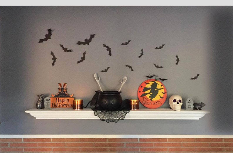 Op de afbeelding: Halloween-thema plankdecoratie met een ketel, schedel en vleermuizen. Inclusief een bord met "Happy Halloween" en een rond bord met een heksensilhouet. De plank is wit en de muur is grijs.
