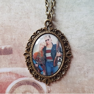 May include: A bronze oval pendant necklace with a cameo featuring a couple wearing sunglasses. The cameo is set in a decorative frame.
