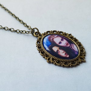 May include: An oval pendant necklace with a bronze-colored chain and frame. The pendant features a portrait of two people with pale skin and dark hair against a blue background. The frame has an ornate, filigree design.