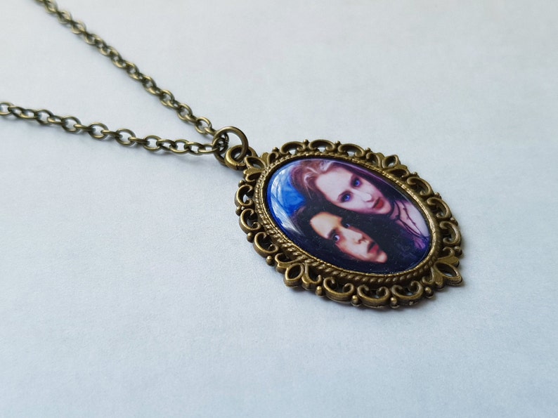 May include: An oval pendant necklace with a bronze-colored chain and frame. The pendant features a portrait of two people with pale skin and dark hair against a blue background. The frame has an ornate, filigree design.