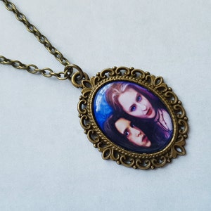 May include: Antique brass-toned oval pendant necklace featuring a portrait of two people with pale skin and dark hair. The pendant has a decorative filigree border and hangs from a matching chain. The background of the portrait is blue.