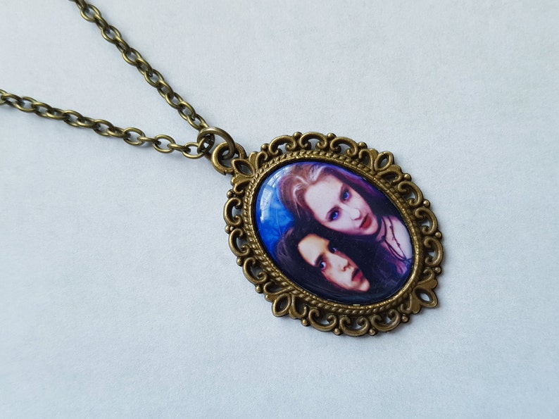 May include: Antique brass-toned oval pendant necklace featuring a portrait of two people with pale skin and dark hair. The pendant has a decorative filigree border and hangs from a matching chain. The background of the portrait is blue.