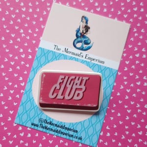 Puede incluir: Pin rectangular con la inscripción blanca "FIGHT CLUB" sobre fondo rojo. El pin está sobre una tarjeta con una ilustración de sirena y el texto "The Mermaid's Emporium". El fondo es rosa con formas de corazón blancas.