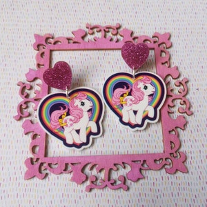 May include: A pair of pink heart-shaped earrings with a rainbow and a My Little Pony illustration. The hearts are covered in glitter.