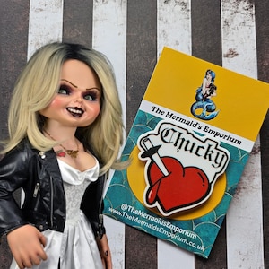 May include: A red heart with a knife through it and the word "Chucky" in white letters. The pin is on a yellow card with the text "The Mermaid's Emporium" and social media information.