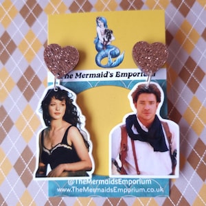 May include: A pair of earrings featuring a heart-shaped charm with glitter. Each charm has a photo of a person on the back. One charm features a woman with dark hair and a black tank top. The other charm features a man with dark hair wearing a blue scarf and a white shirt. The text "@TheMermaidsEmporium" and "www.TheMermaidsEmporium.co.uk" is printed on the card.