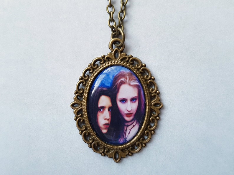 May include: An antique brass-toned pendant necklace featuring an oval cameo with a portrait of two people. The cameo is set within a decorative filigree frame and hangs from a matching chain. The portrait has a blue background.