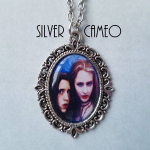 May include: Silver cameo pendant with an oval frame and a chain. The cameo features a portrait of two people with pale skin, dark hair, and red lips. The text "SILVER CAMEO" is above the pendant.