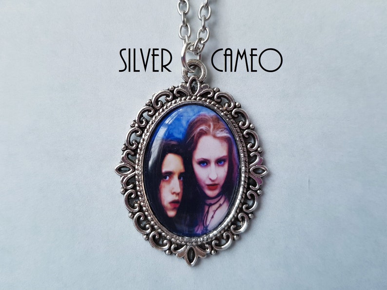 May include: Silver cameo pendant with an oval frame and a chain. The cameo features a portrait of two people with pale skin, dark hair, and red lips. The text "SILVER CAMEO" is above the pendant.