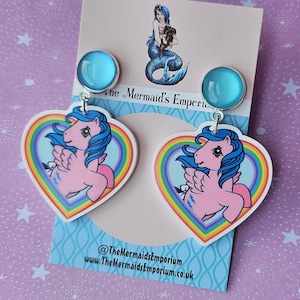May include: A pair of earrings featuring a pink pony design within a rainbow heart. The earrings have a light blue stud and are attached to a white backing. The Mermaid's Emporium is printed on the backing.