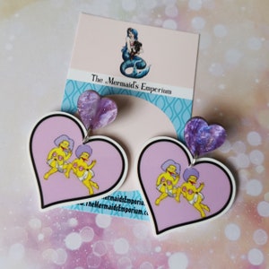 May include: A pair of heart-shaped earrings with a purple glitter design. Each earring features a pink heart with a cartoon image of two yellow characters with blue hair. The text "The Mermaid's Emporium" is visible on a card behind the earrings.