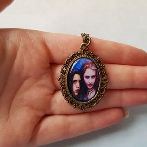 May include: Oval pendant with a bronze-colored filigree frame. The pendant features a portrait of two women with pale skin and dark hair against a blue background. The pendant is suspended from a matching bronze chain.