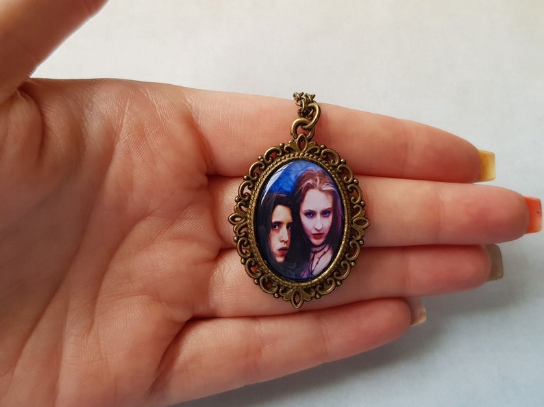 May include: Oval pendant with a bronze-colored filigree frame. The pendant features a portrait of two women with pale skin and dark hair against a blue background. The pendant is suspended from a matching bronze chain.