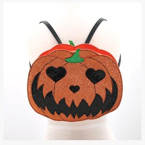 May include: Orange pumpkin-shaped backpack with black glitter and black heart eyes and a toothy grin. The backpack has black adjustable straps.