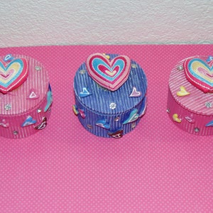 May include: Three small round boxes with heart-shaped lids. The boxes are decorated with hearts and rhinestones. The boxes are pink, blue, and white.