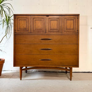 May include: A tall wooden dresser featuring a top section with four square-paneled cabinet doors and three lower drawers. Each drawer is equipped with a horizontal, almond-shaped recessed handle. The piece stands on four tapered wooden legs.