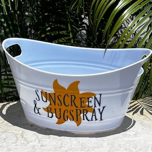 May include: White oval metal tub with light blue interior and handles. The tub has the words "SUNSCREEN & BUGSPRAY" in black and orange lettering. The tub is sitting outside with green foliage in the background.