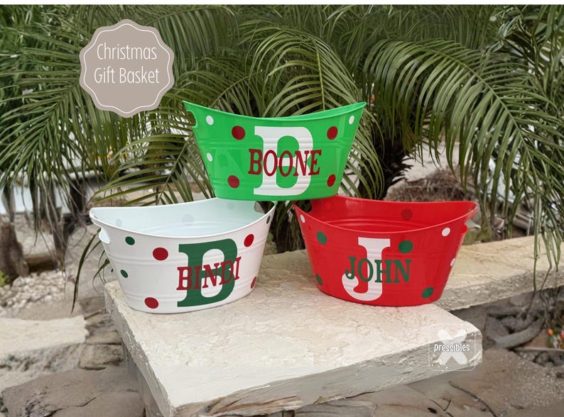 Personalized Christmas Gift Basket Custom Family Gift Holiday Goodie Basket Santa Sack Custom Reusable Holiday Cheer Bucket Name Basket May include: Three personalized Christmas tubs in green, white, and red. Each tub features a large initial and name, such as 'Boone', 'BinDi', and 'John', in a festive green font. The tubs are decorated with red and green polka dots on a white or colored background. These decorative storage containers are perfect for holiday gift-giving or organization.