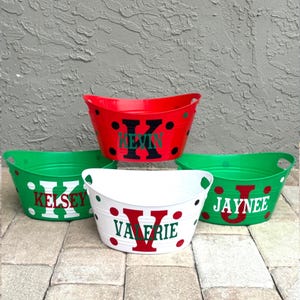 May include: Four personalised oval tubs in red, green, and white. Each tub has a large initial and name in a contrasting colour. The red tub has black polka dots. The tubs are on a stone surface.