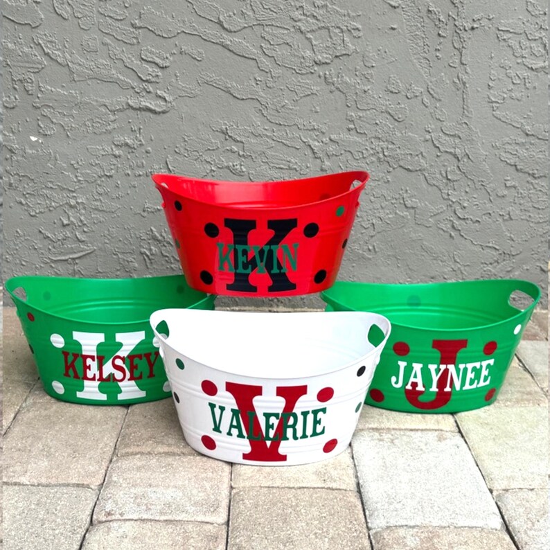 May include: Four personalised oval tubs in red, green, and white. Each tub has a large initial and name in a contrasting colour. The red tub has black polka dots. The tubs are on a stone surface.
