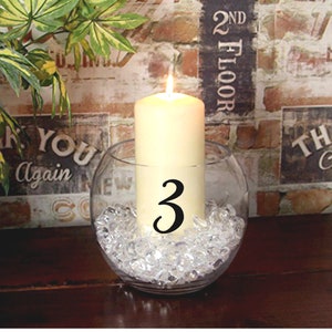 May include: A white candle with a black number 3 on it sits in a clear glass vase filled with clear glass stones. The vase is on a wooden table in front of a brick wall.