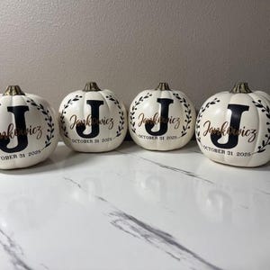 May include: Four white ceramic pumpkins with gold stems. Each pumpkin has a black letter "J" and the name "Jankowicz" in gold script, surrounded by a wreath. The date "October 31 2025" is also displayed.