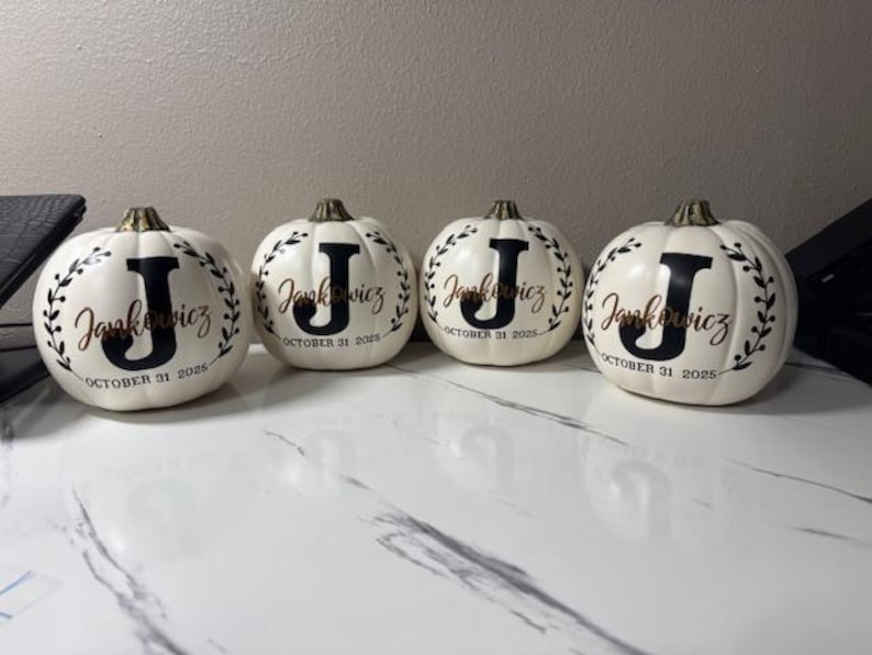 May include: Four white ceramic pumpkins with gold stems. Each pumpkin has a black letter "J" and the name "Jankowicz" in gold script, surrounded by a wreath. The date "October 31 2025" is also displayed.