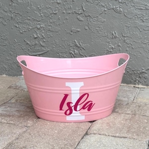 May include: A pink metal tub with handles, featuring the name "Isla" in a cursive font with a white "I" initial. The tub has a rounded shape and is sitting on a stone surface.