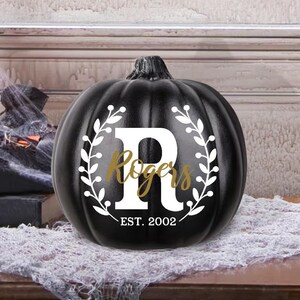 May include: A black pumpkin with a white floral wreath design and the letter "R". The word "Rogers" is in gold script, with "EST. 2002" below. A decorative item for Halloween or fall.