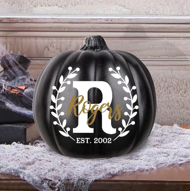 May include: A black pumpkin with a white floral wreath design and the letter "R". The word "Rogers" is in gold script, with "EST. 2002" below. A decorative item for Halloween or fall.