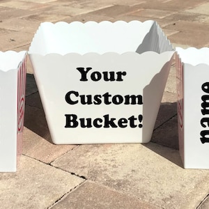 May include: A set of white popcorn buckets with red and white striped sides. The buckets have a scalloped top edge and the word "name" is printed in black on four of the buckets. The center bucket has the text "Your Custom Bucket!"