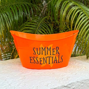 May include: Bright orange metal tub with curved handles. The words "SUMMER ESSENTIALS" are printed on the front in dark blue, with a yellow sun graphic. The tub is sitting on a white surface, with green foliage in the background.