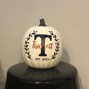 May include: Cream-colored pumpkin with a black stem, featuring a black letter "T" and the name "Tharrett" in gold script, encircled by a black leaf wreath. The text "EST. 2023" is below the name. The pumpkin is displayed on a black surface.
