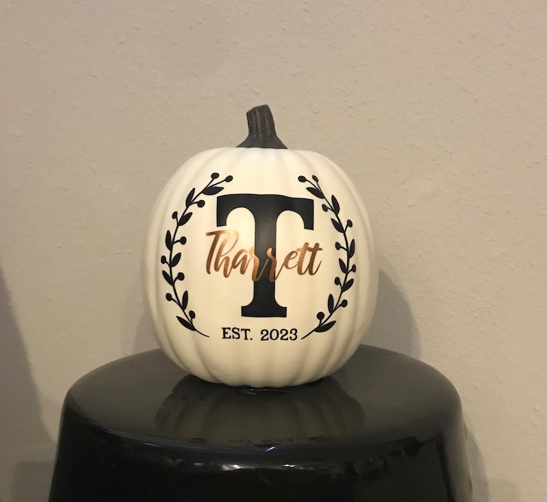 May include: Cream-colored pumpkin with a black stem, featuring a black letter "T" and the name "Tharrett" in gold script, encircled by a black leaf wreath. The text "EST. 2023" is below the name. The pumpkin is displayed on a black surface.