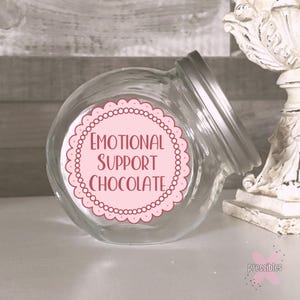 May include: A clear glass jar with a silver lid, tilted slightly. A pink label with a scalloped edge and the words "EMOTIONAL SUPPORT CHOCOLATE" in a serif font is affixed to the jar. The jar is on a white surface.